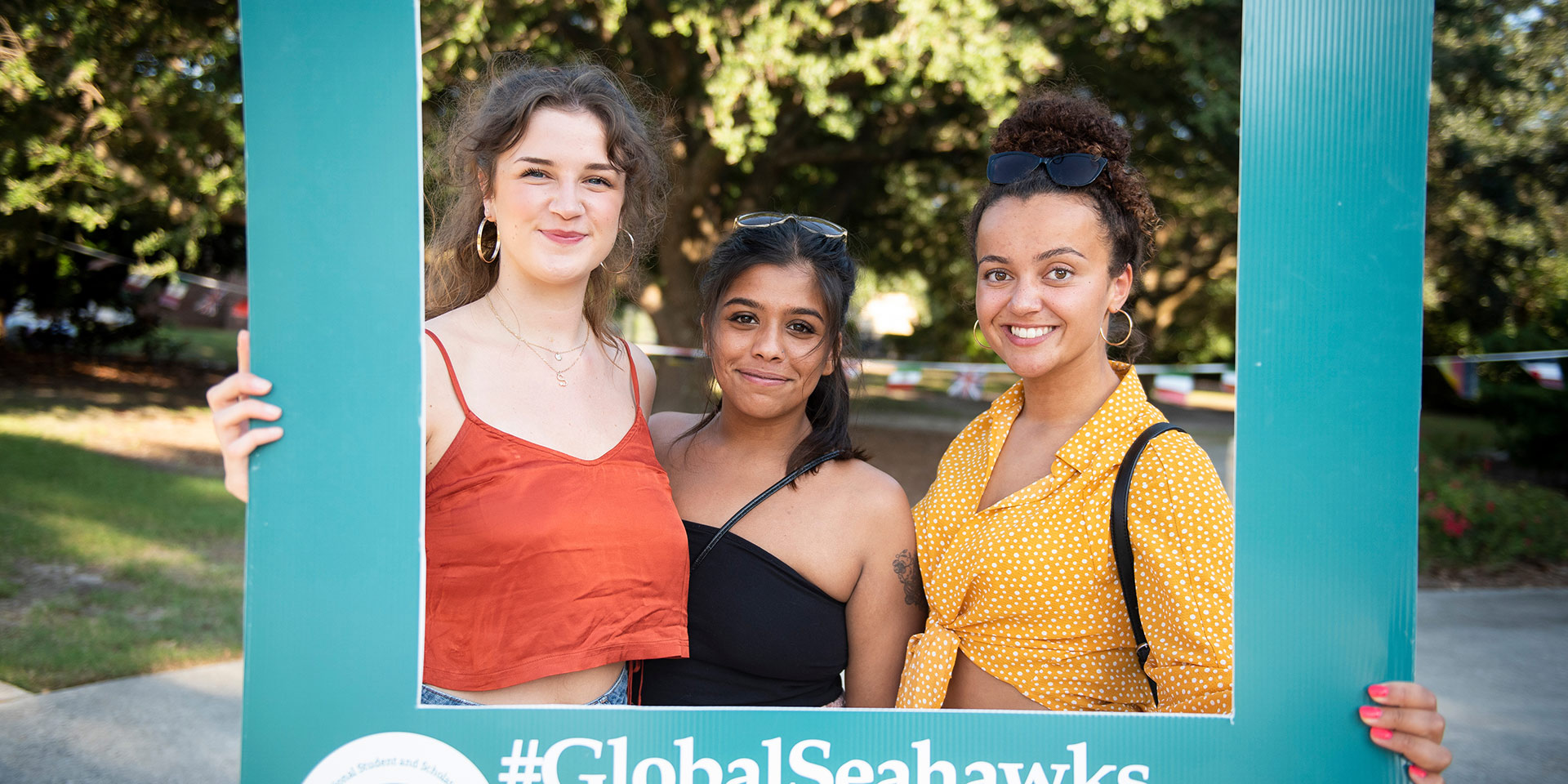 Three students hold a sign framing their faces that says #GlobalSeahawks