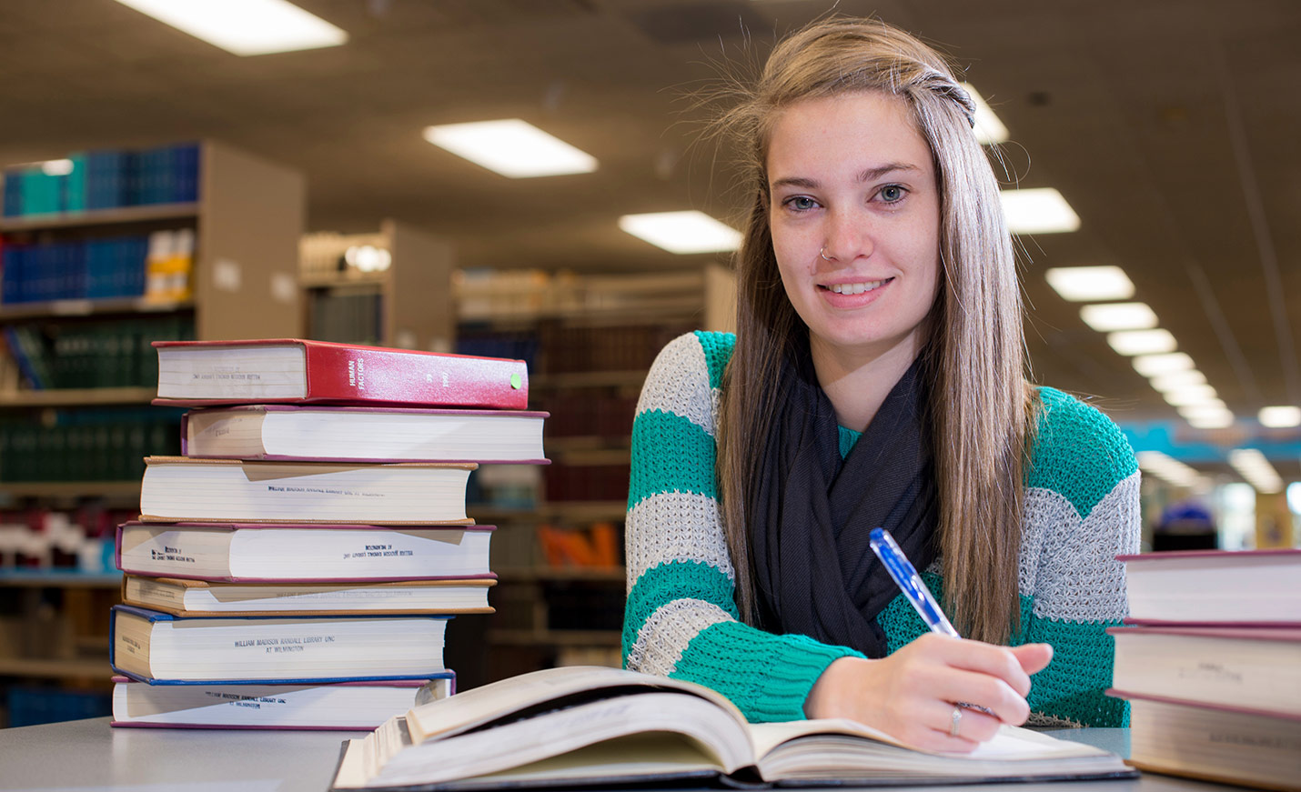 A student holding a pen sits at a table next to a stack of books