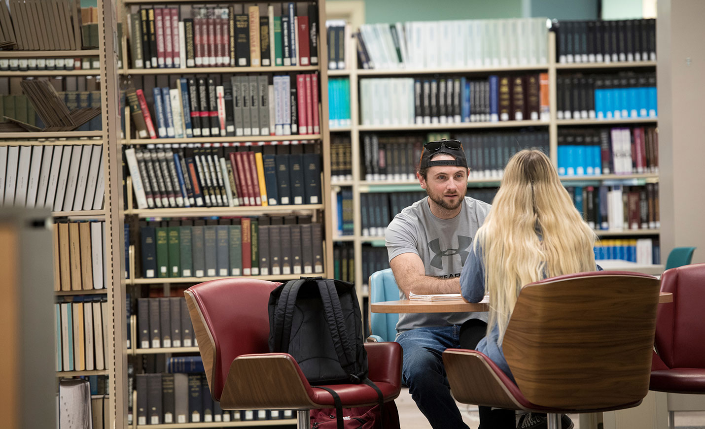 Two students sit across a table from each other in the stacks of the library