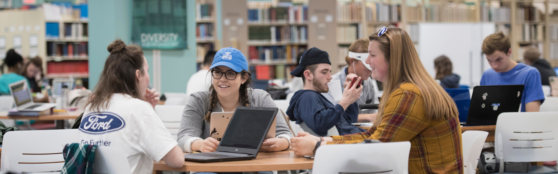 Students sit at table in library while studying on their laptops