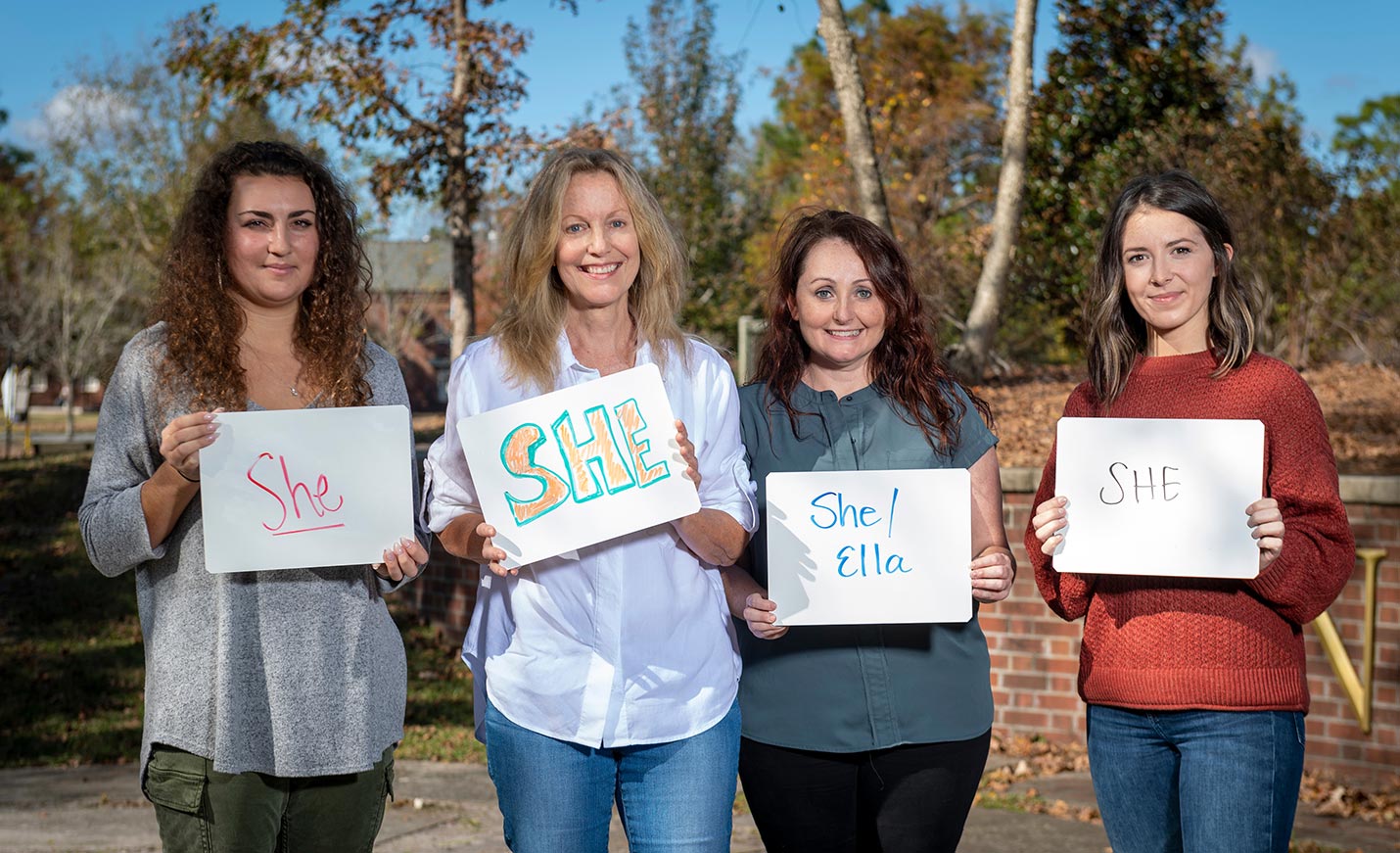 Four women holding signs indicating their preferred pronoun is She; one added the Spanish word ella