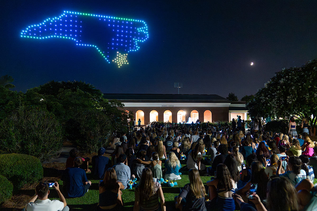 Drones form an image of North Carolina with a star marking Wilmington in the night sky above campus as a crowd watches