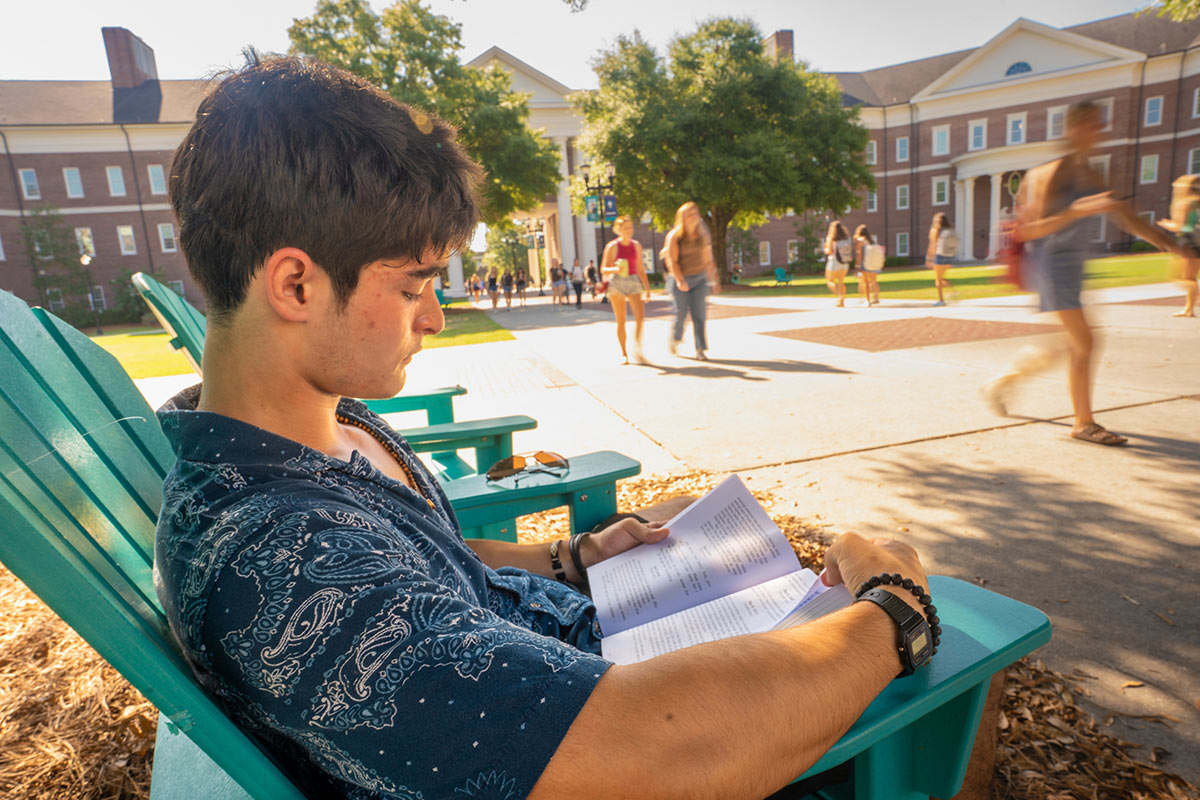 A student sits outside reading as others walk past on campus