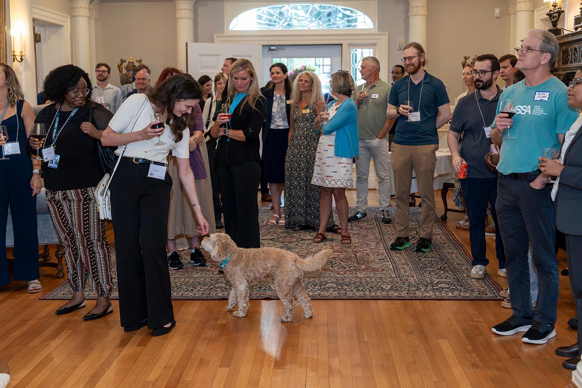 A person in a group congregating in the foyer of Kenan House leans over to pet Toby, a dog.