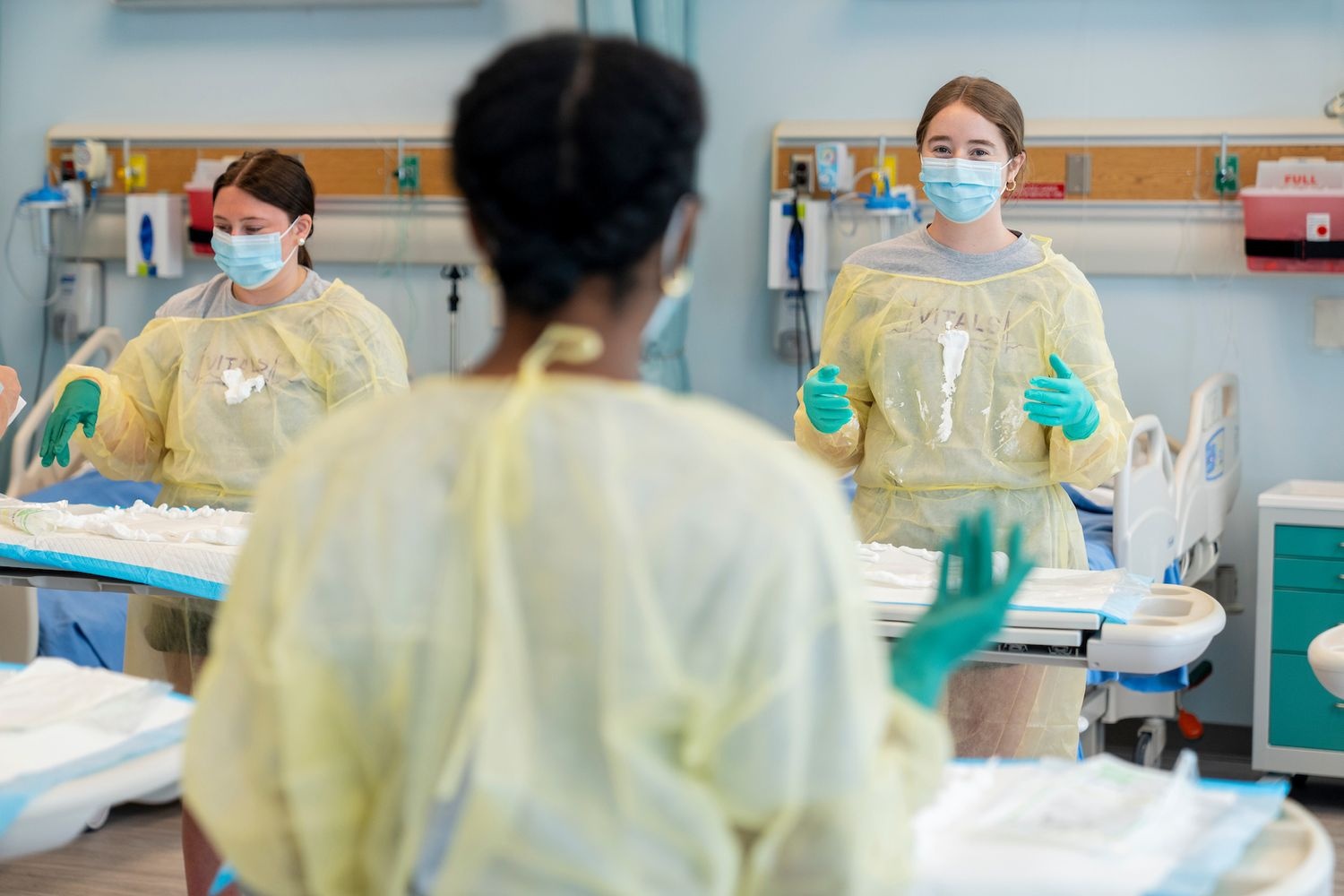 Nursing students wearing protective gowns and gloves in a classroom lab with equipment.