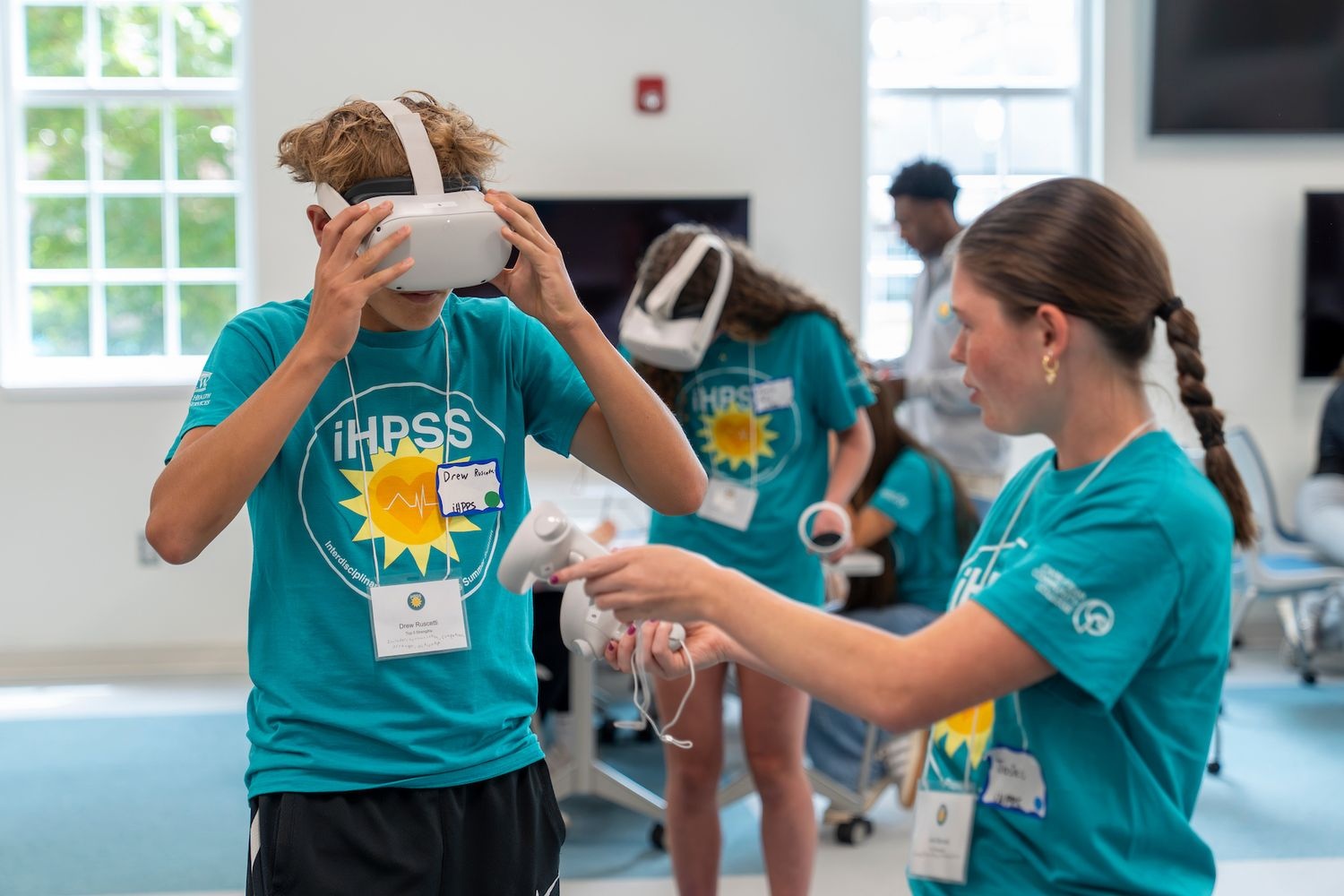 An iHPSS summer camper helps another camper put on a vr headset that features a healthcare simulation.