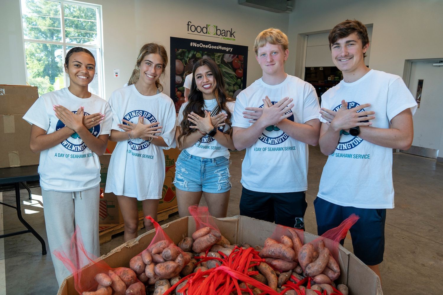 Group holding up wings up hand symbol in while standing behind bags of potatoes