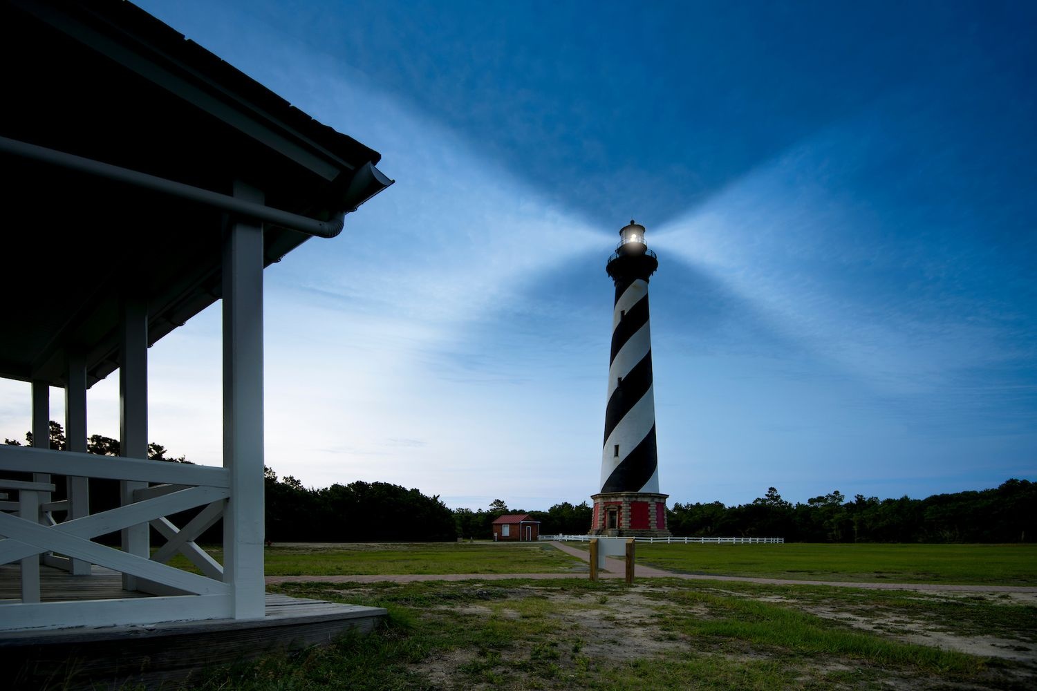 Spiral-striped lighthouse shining at twilight, with a small building nearby and a porch in the foreground