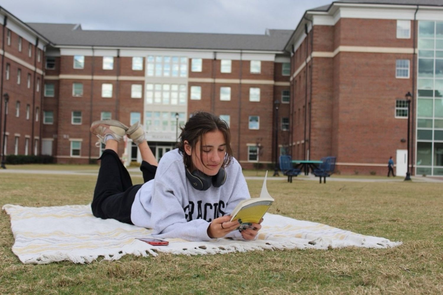 A student lies on a blanket on a grassy campus lawn, reading a book with headphones around her neck. A large brick residence hall stands in the background.