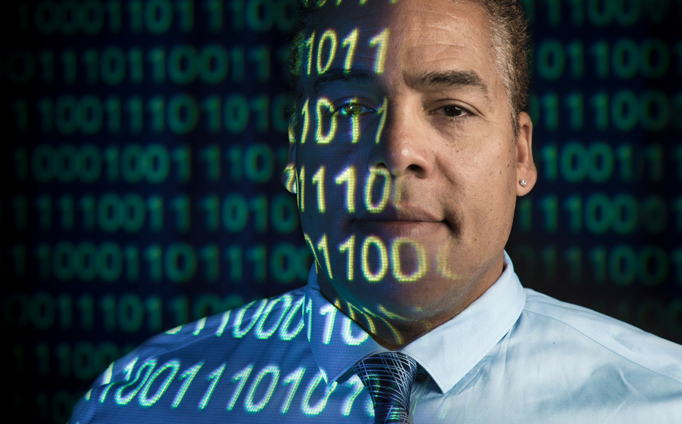 A professor looks at the camera with 1’s and 0’s projected onto his face
